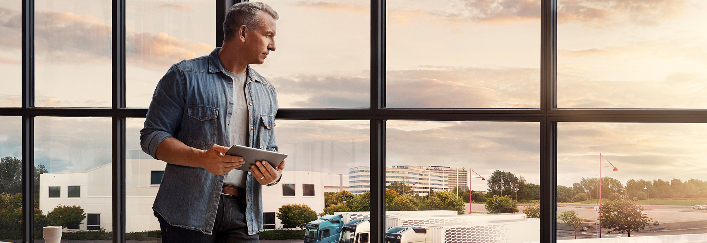 Човек кој држи таблет стои покрај прозорецот и гледа надолу кон возниот парк од камиони